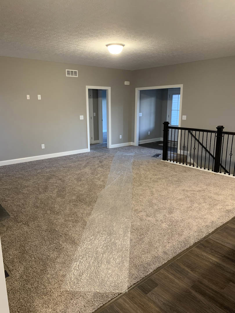 Carpeted room with white trim, staircase featuring black metal railing, hallway with doorway and wall-mounted light switch, neutral walls and ceiling