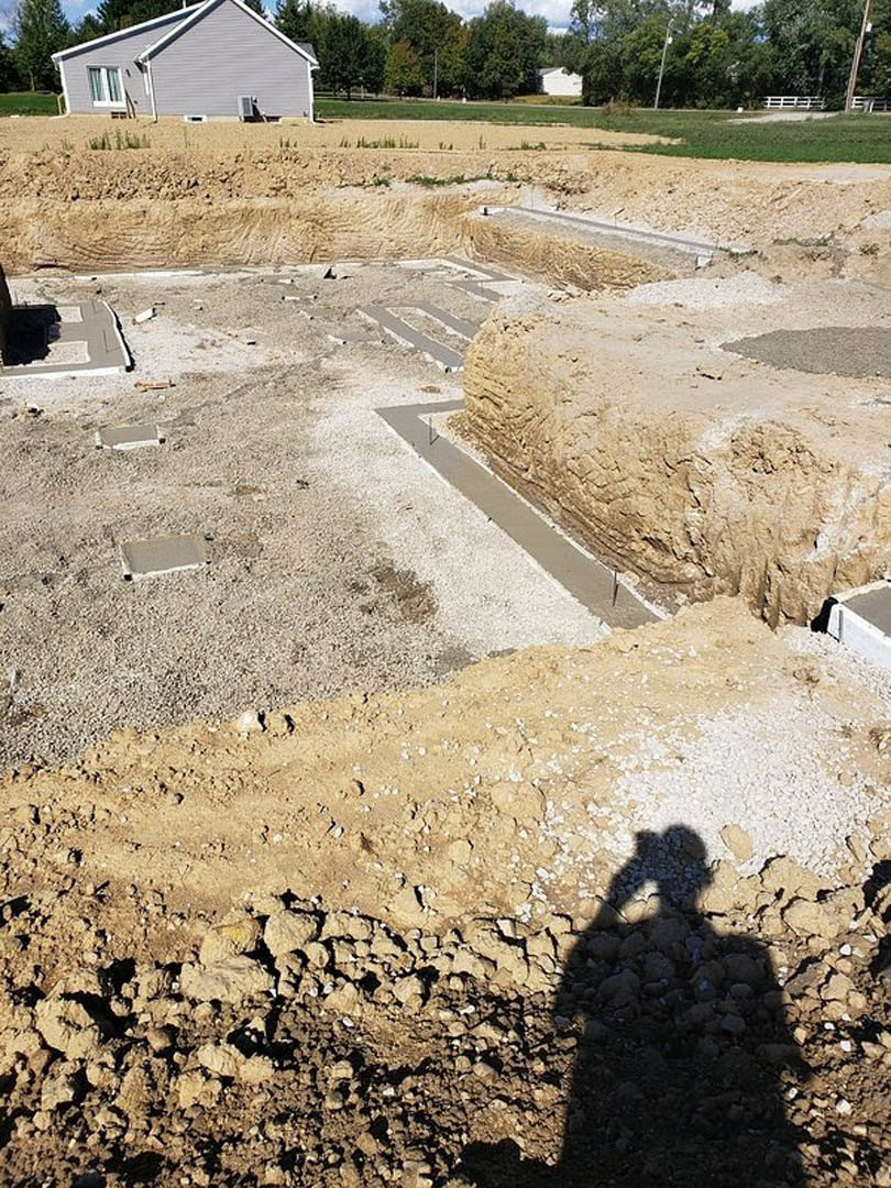 Grey house with white trim under construction, concrete path beside a stacked rock wall, grassy field and trees in background, shadow of person visible on ground