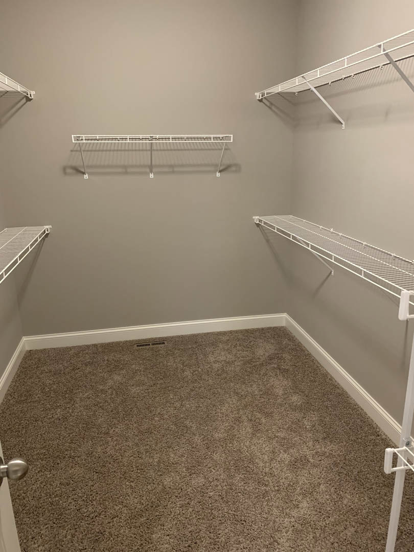 Empty walk-in closet with white shelving, metal hanging bars, carpeted floor, wall vent, and white baseboards.