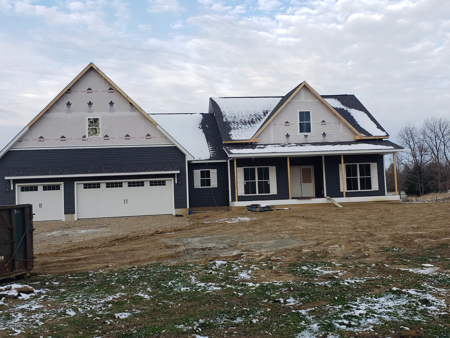 Partially built house with white-framed windows, triangle roof, and exposed dirt patch in front, surrounded by snow and trees in the background; metal box with chain visible near