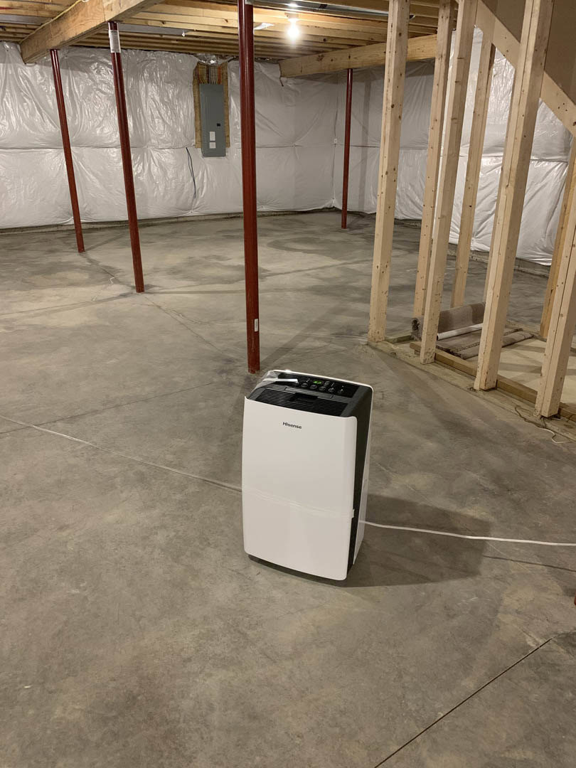White portable air conditioner with black handle positioned on concrete basement floor, exposed wooden ceiling beams, white door with black square accent, neutral walls.