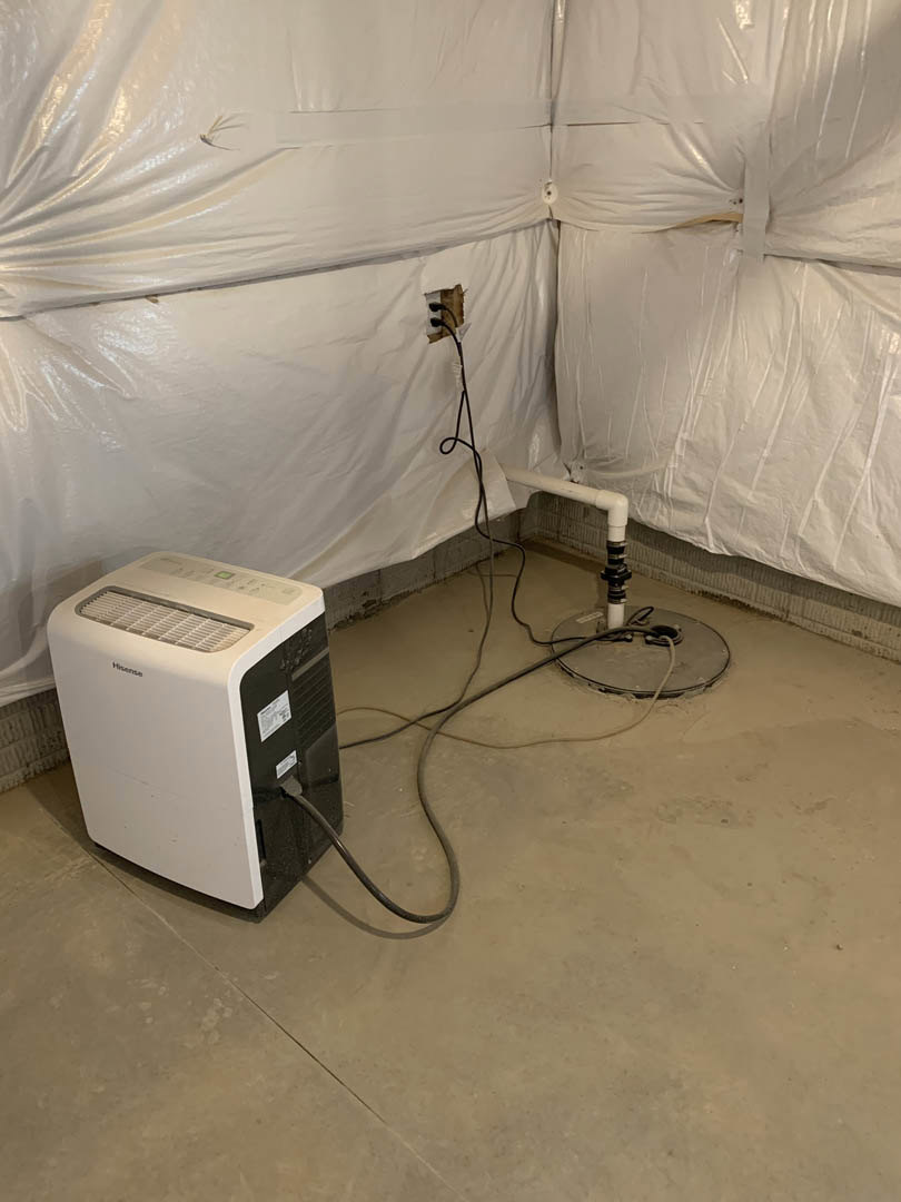 White electrical box with exposed wires mounted on a round base, surrounded by plastic sheeting in a room with unfinished floors and walls.
