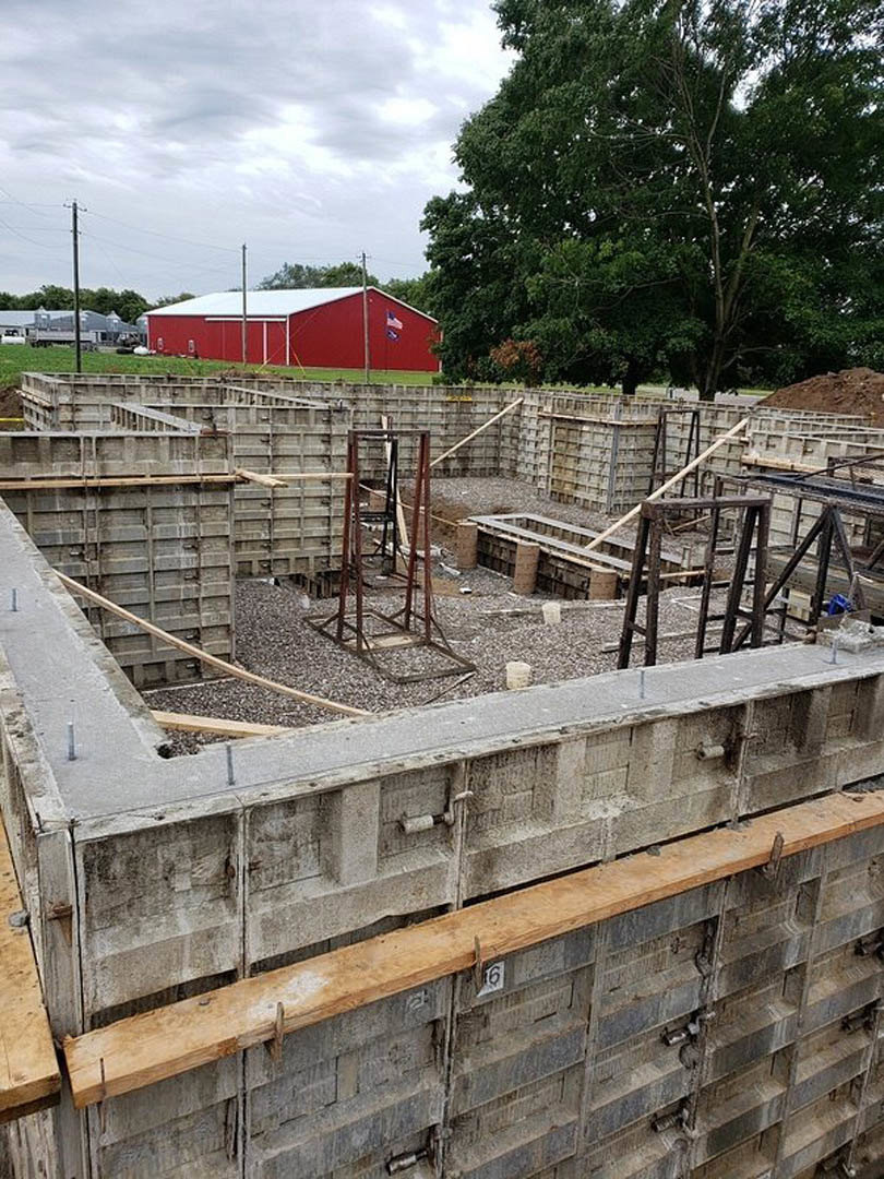 Partially built house with exposed metal framing and concrete foundation, red barn with white roof in background, surrounded by mature trees and grassy outdoor area.