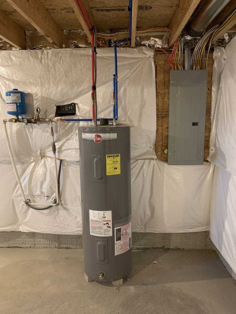 Water heater installed against a white wall in a utility room, grey door with square cutout, blue container with white label, yellow caution sign, and white plastic bag with handle