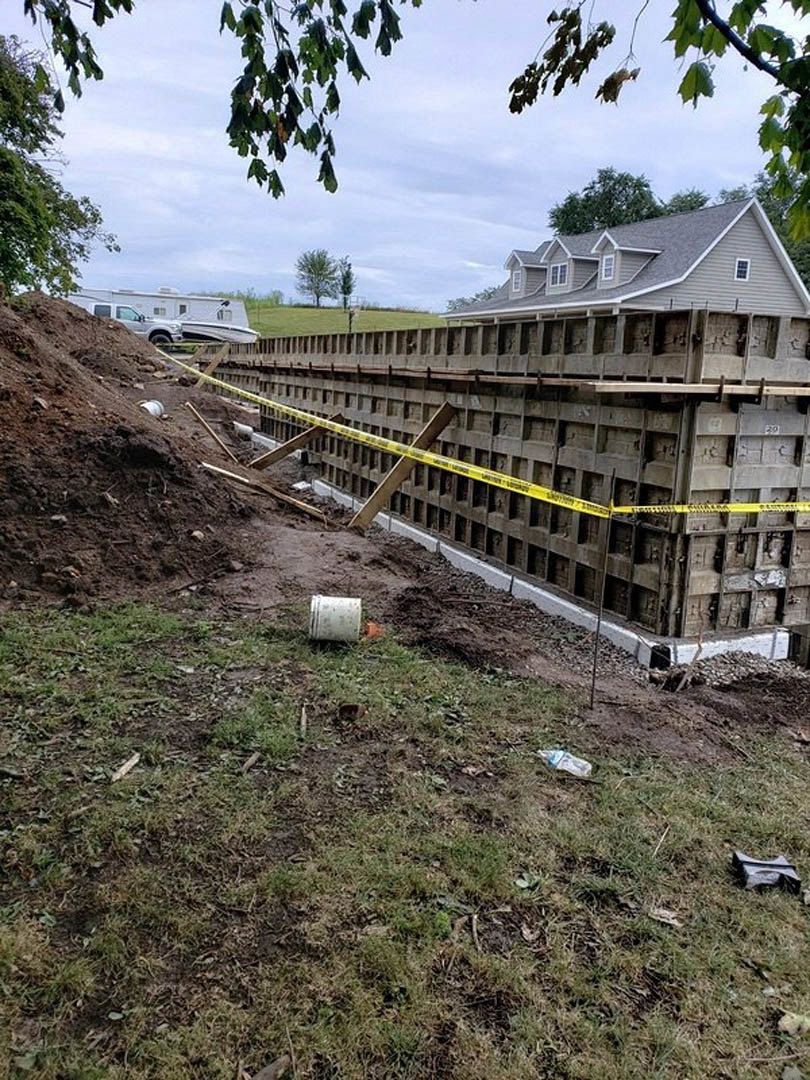 Partially built house with exposed framing, yellow caution tape stretched across the site, white bucket on dirt, grey construction material in grass, leafy tree nearby, cloudy sky