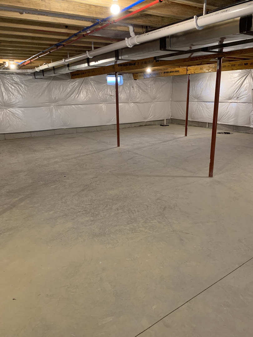 Basement room with concrete floor, exposed ceiling pipes, steel and wooden support poles, white plastic sheets covering walls