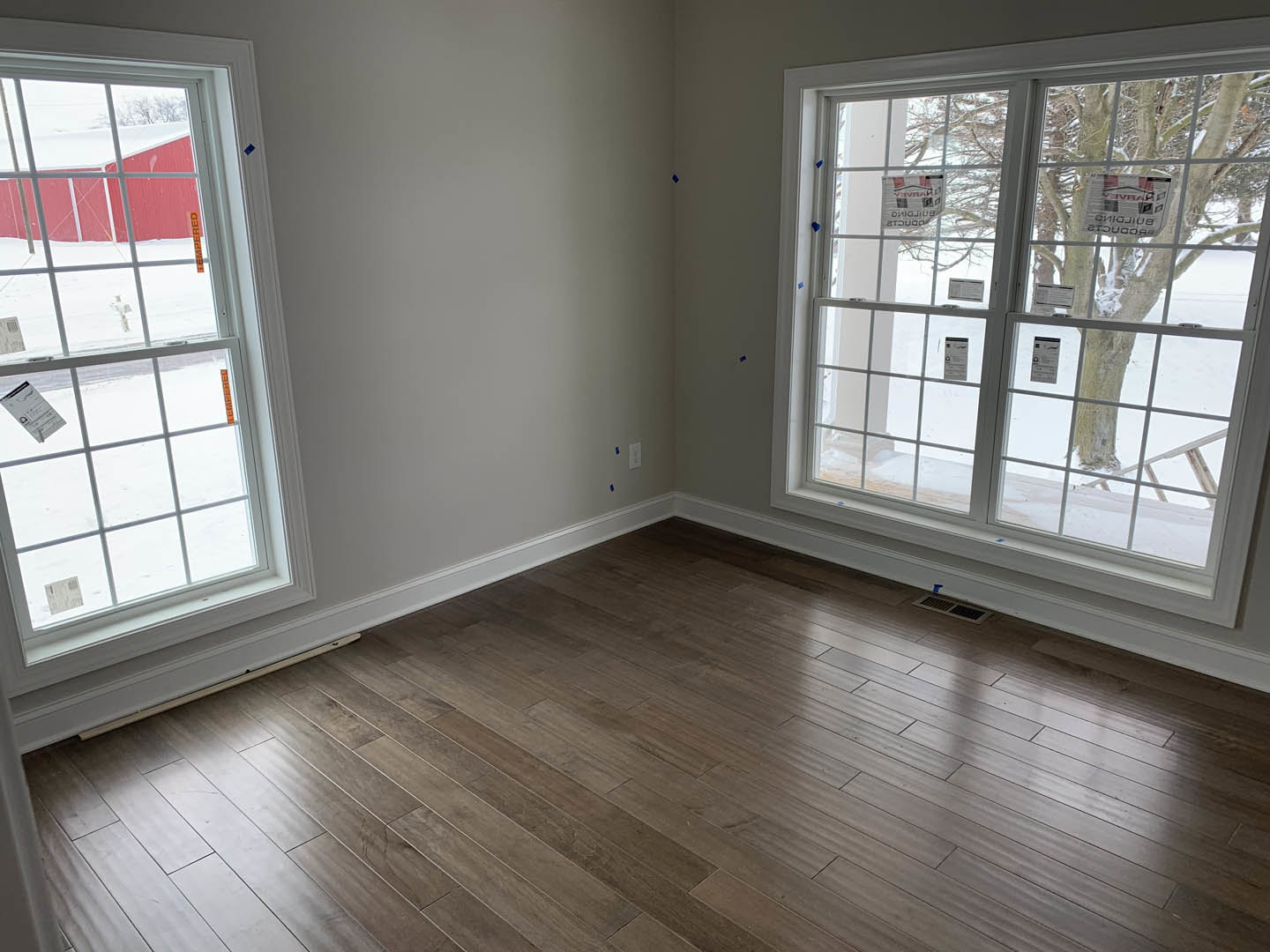 Sunlit room featuring large windows with views of a snowy landscape and red building, wood flooring, white walls, and a window displaying a sign.