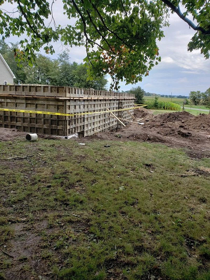 Partially built residential structure with exposed framing, yellow caution tape, stacks of crates, dirt lot, green tree and grass in foreground, cloudy sky overhead