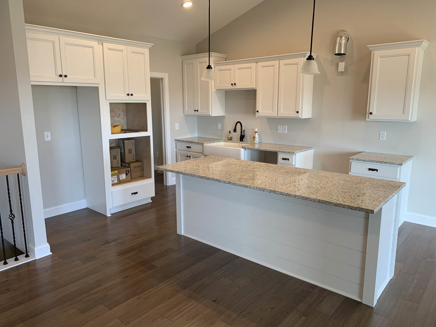 Spacious kitchen featuring a large central island with stone countertop, wood flooring, white cabinetry, tile backsplash, stainless steel sink, and modern metal railing visible in