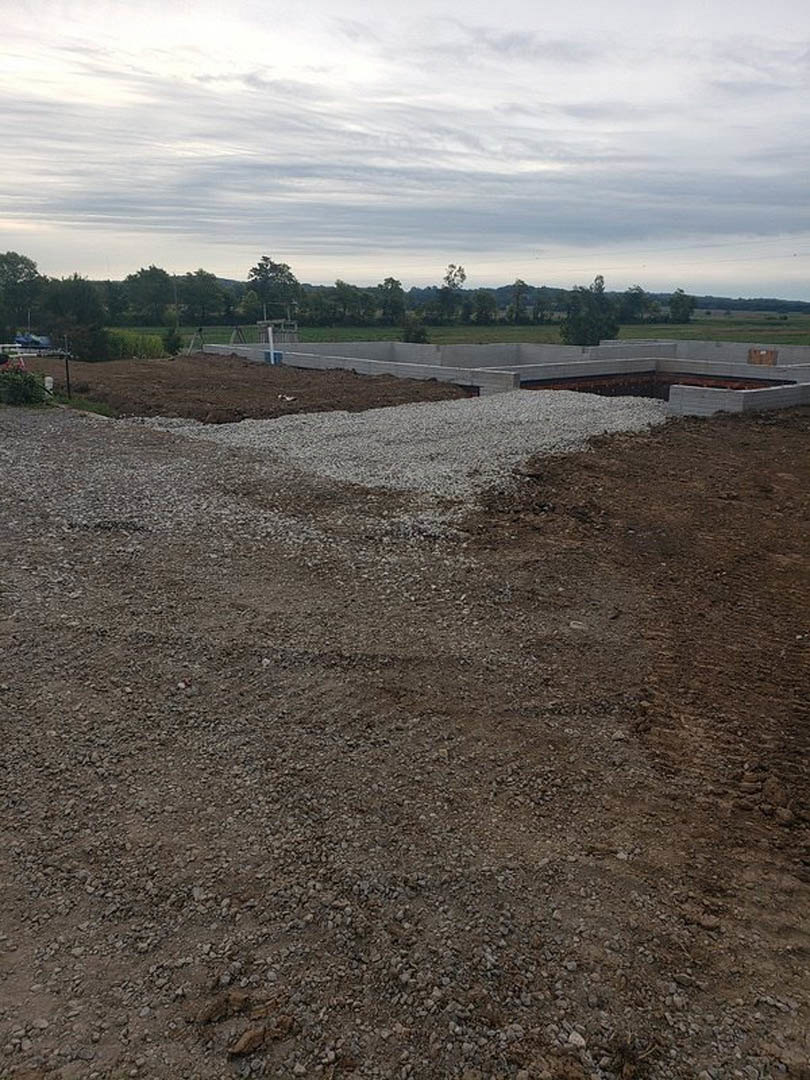 Concrete foundation and retaining wall on a dirt and gravel construction site, bush nearby, cloudy sky overhead