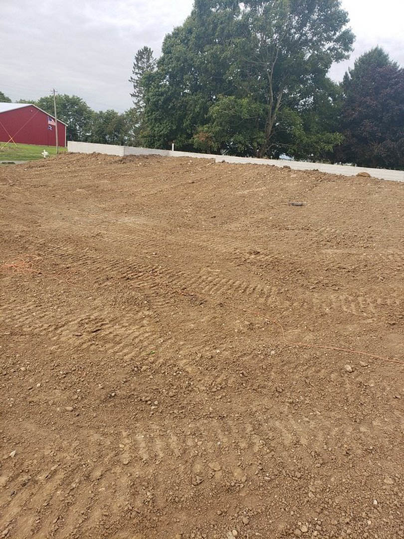 Dirt field with tire tracks bordered by leafy trees, red barn with flag in background, rural landscape under open sky
