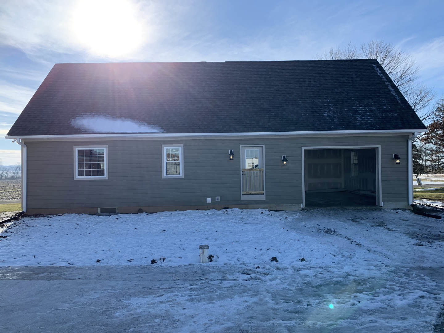 Two-story house with gray siding, white trim, and multiple windows, snow covering the lawn and driveway, overcast winter sky in the background