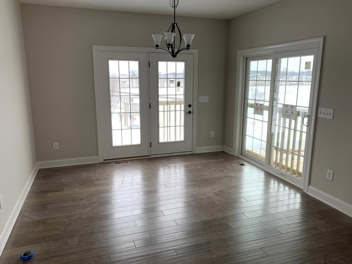 Wood flooring in a spacious room, ornate chandelier hanging from ceiling, glass-paneled door with a sign, window letting in natural light, roll of blue tape near entryway, white