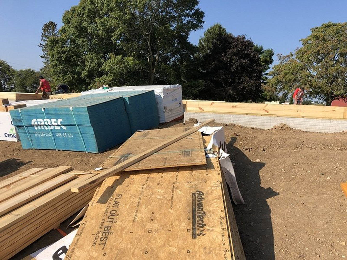 Framed house under construction with exposed plywood, stacked lumber, soil foundation, and a worker in a red shirt near a blue sign