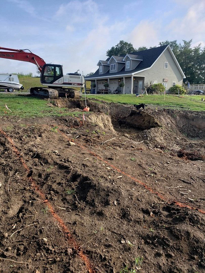 Partially built house with attached garage, exposed framing, bulldozer parked on dirt lot, boat on trailer beside driveway, dog running across soil, white vehicle with black