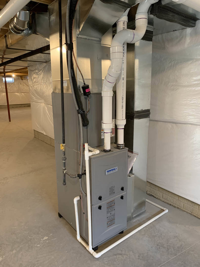 Grey HVAC unit with white PVC pipes and metal ductwork installed against an unfinished wall with exposed electrical wiring and steel framing.