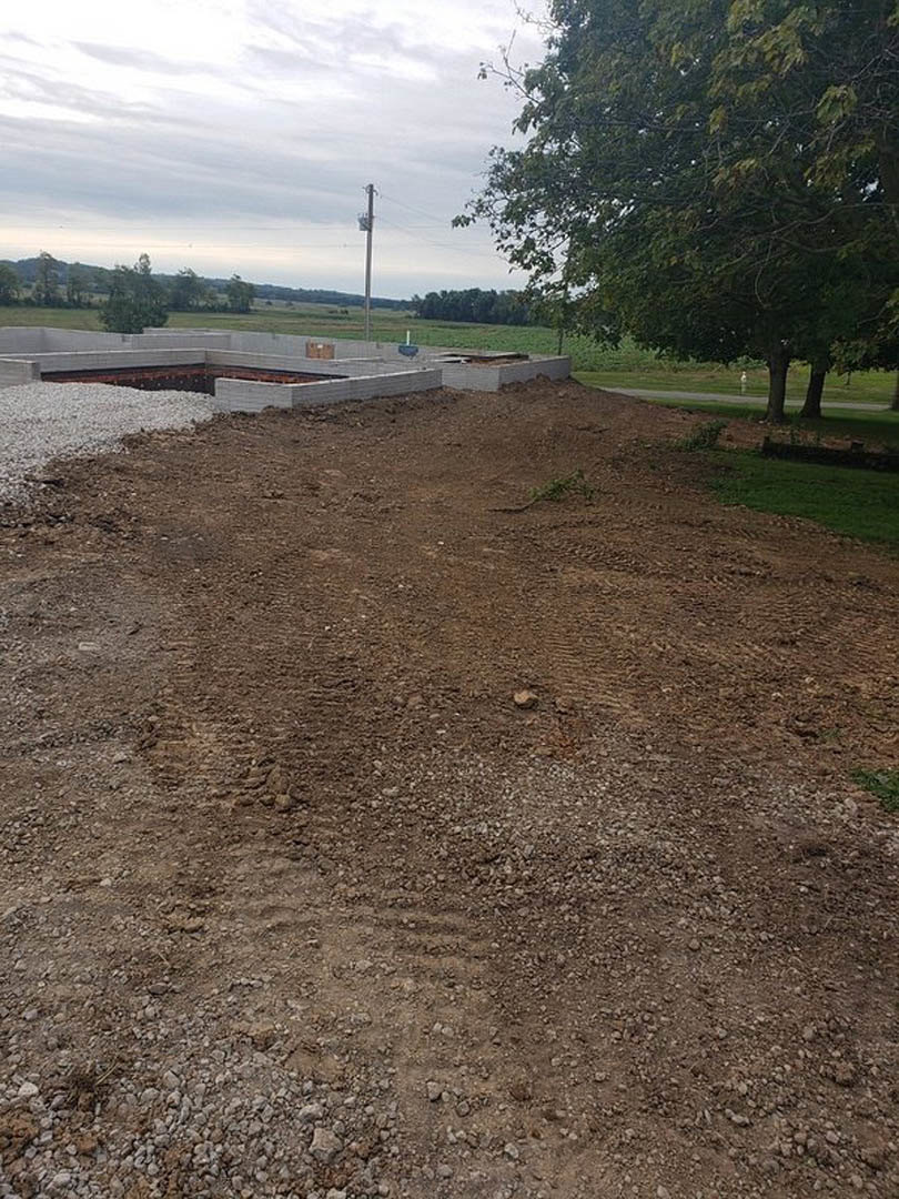 Dirt lot with exposed building foundation, mature tree, gravel piles, and brick wall under partly cloudy sky
