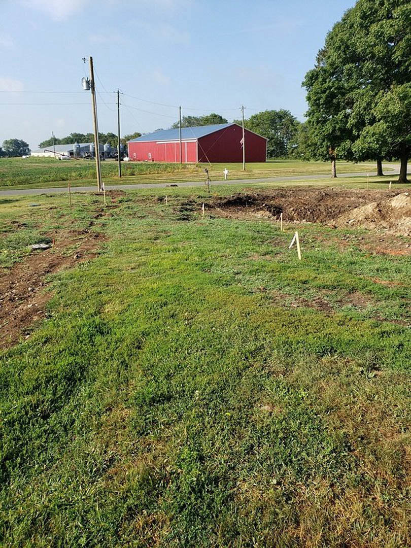 Grassy field with scattered dirt and poles, leafy tree, red barn with blue roof in the background, blue sky with clouds overhead