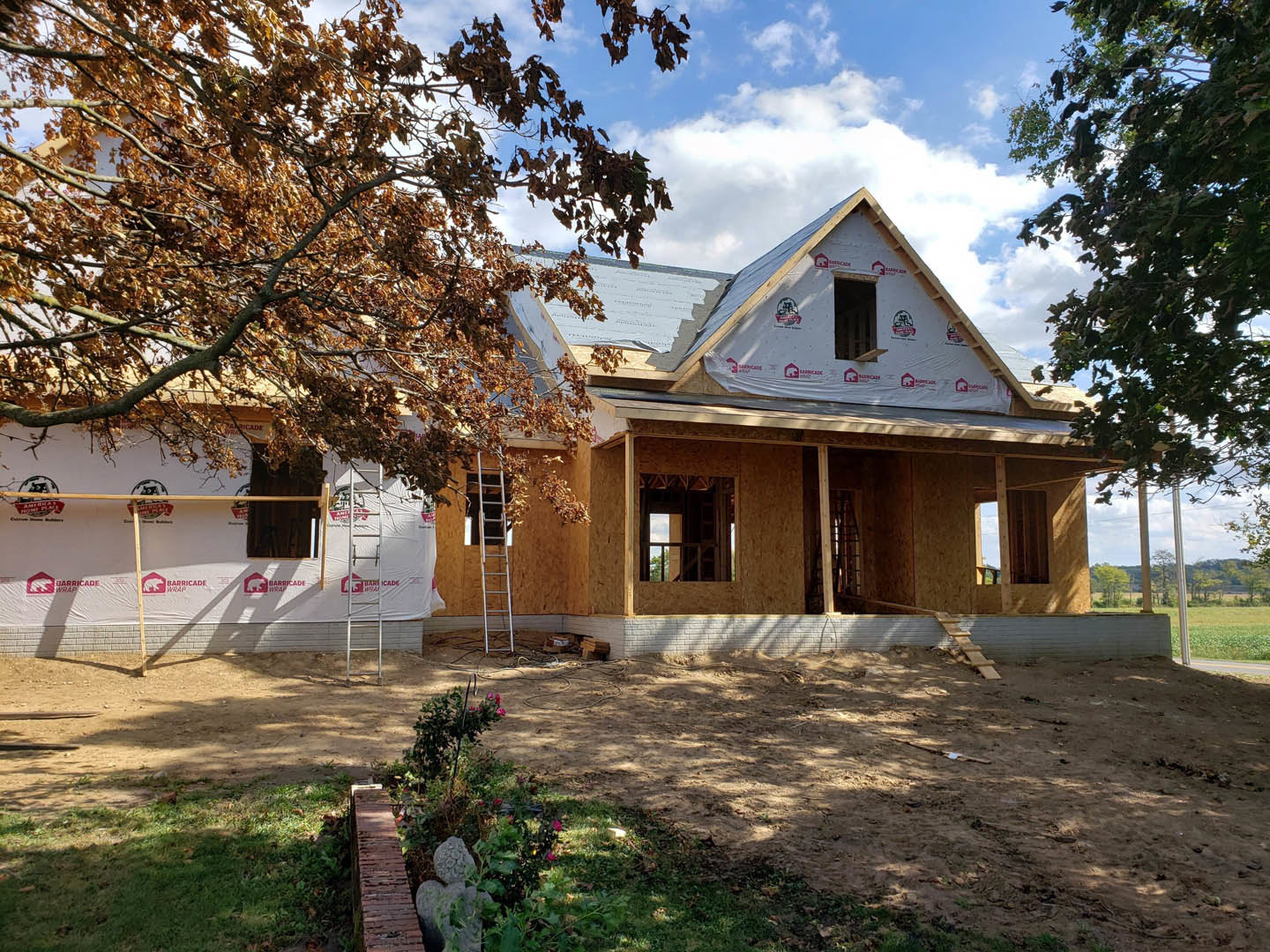 Two-story house under construction with exposed framing, white window frames, dirt yard, brick wall, wooden fence, and surrounding trees
