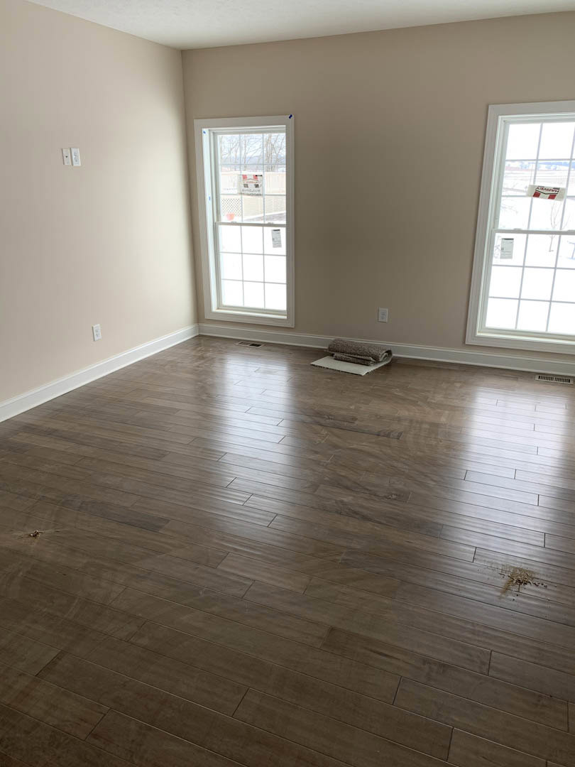 Hardwood floor with area rug, two windows with white trim, neutral plaster walls, roll of carpet resting on protective mat