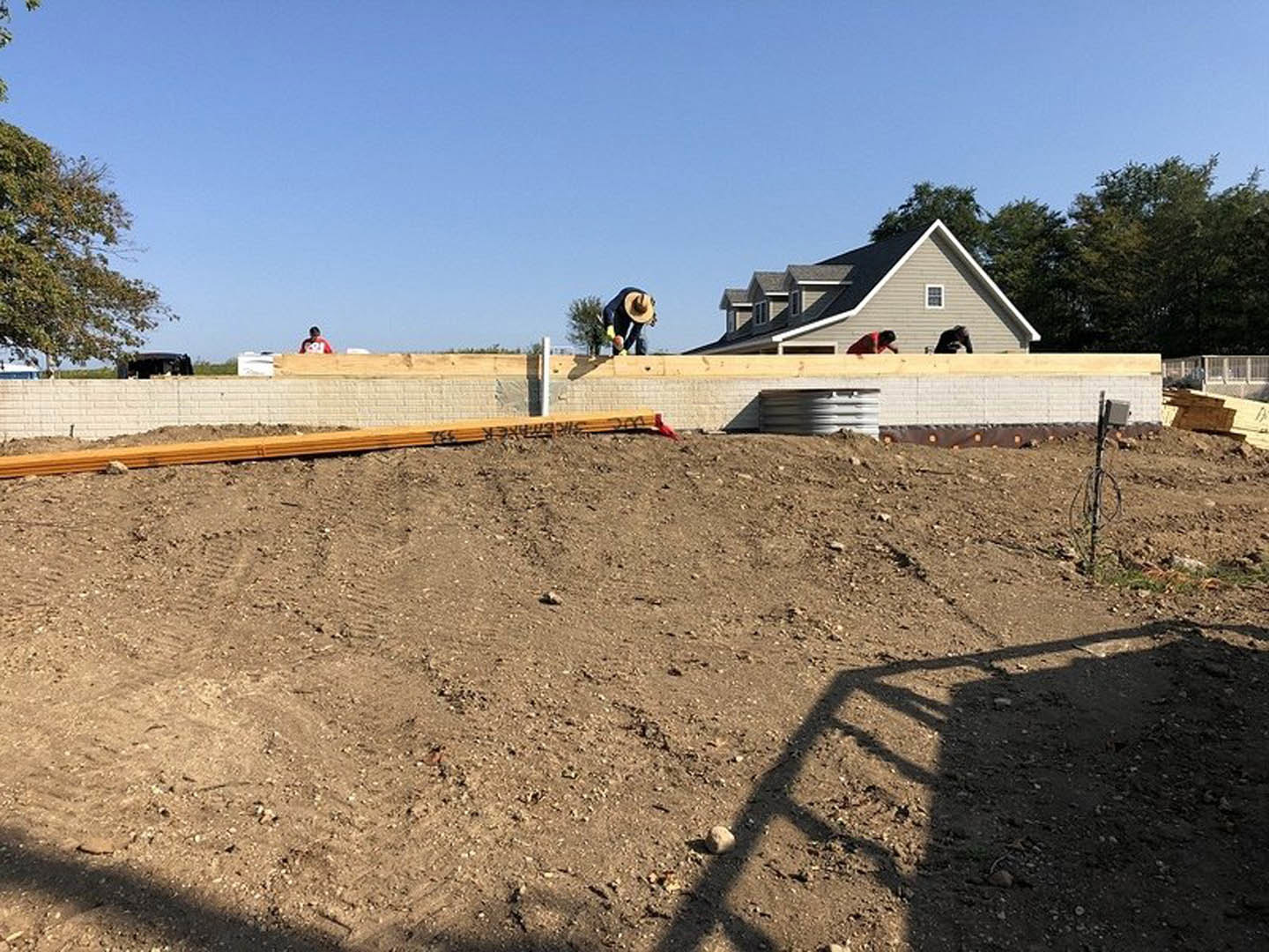 Partially built house with exposed wooden framing, roof structure, and fenced construction site; dirt ground with ladder shadow, nearby soil mound, and person in hat standing near