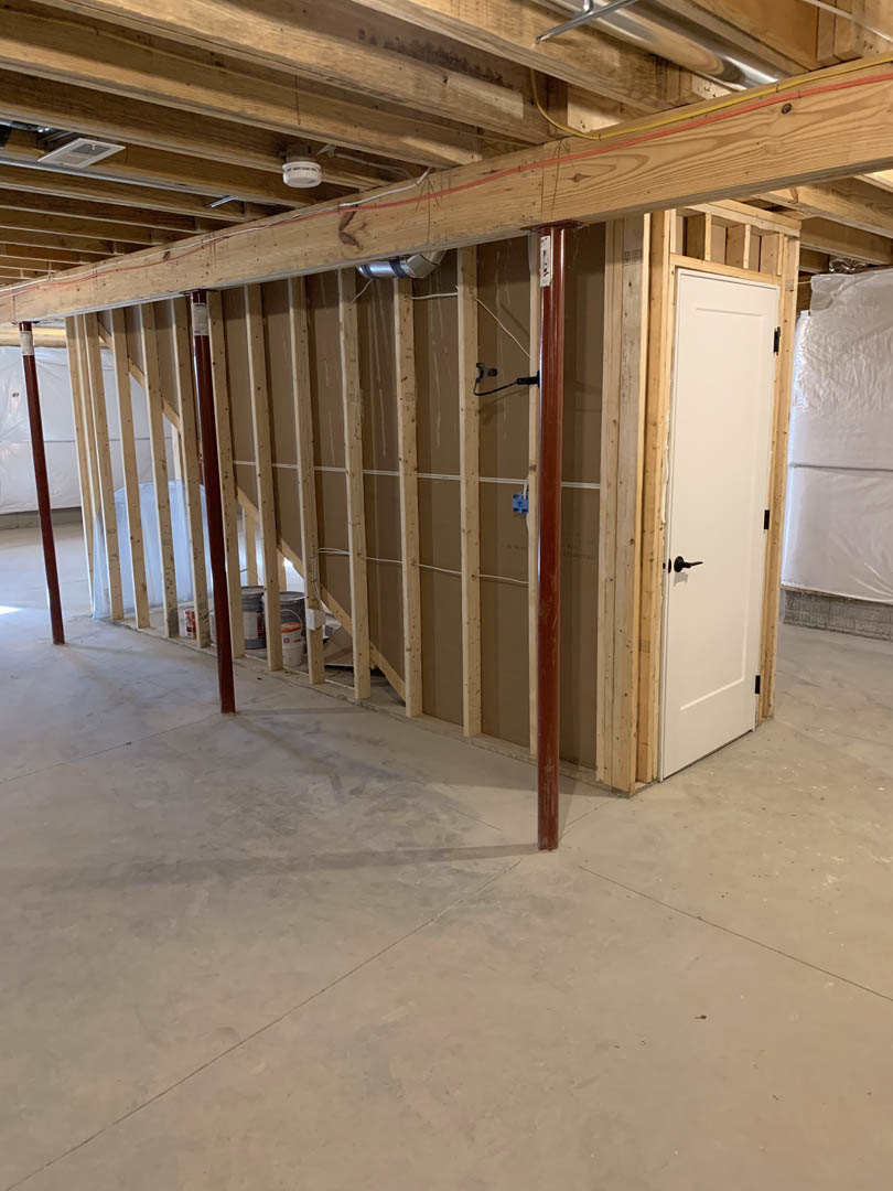 Unfinished room with exposed wood beams, red pipes, concrete floor, white door with black handle, and plastered wall