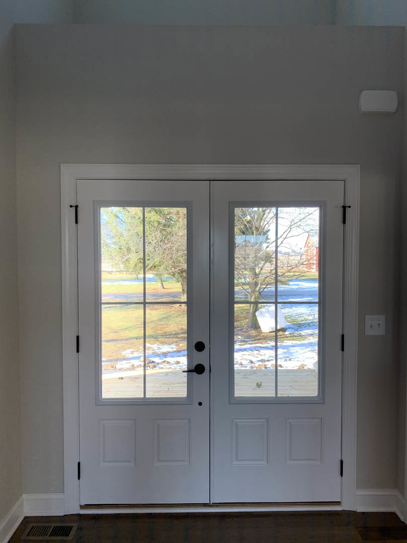 Double doors with glass panes framed by white trim, a light switch on the adjacent wall, window overlooking grassy field, white rectangular object with black border near entry
