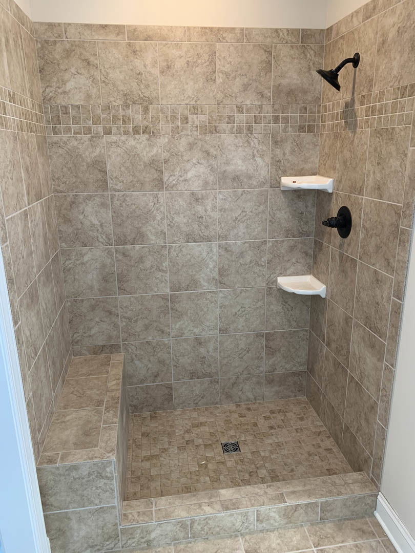 Walk-in shower with marble tile walls, built-in bench, two white corner shelves, black metal showerhead, and floor drain