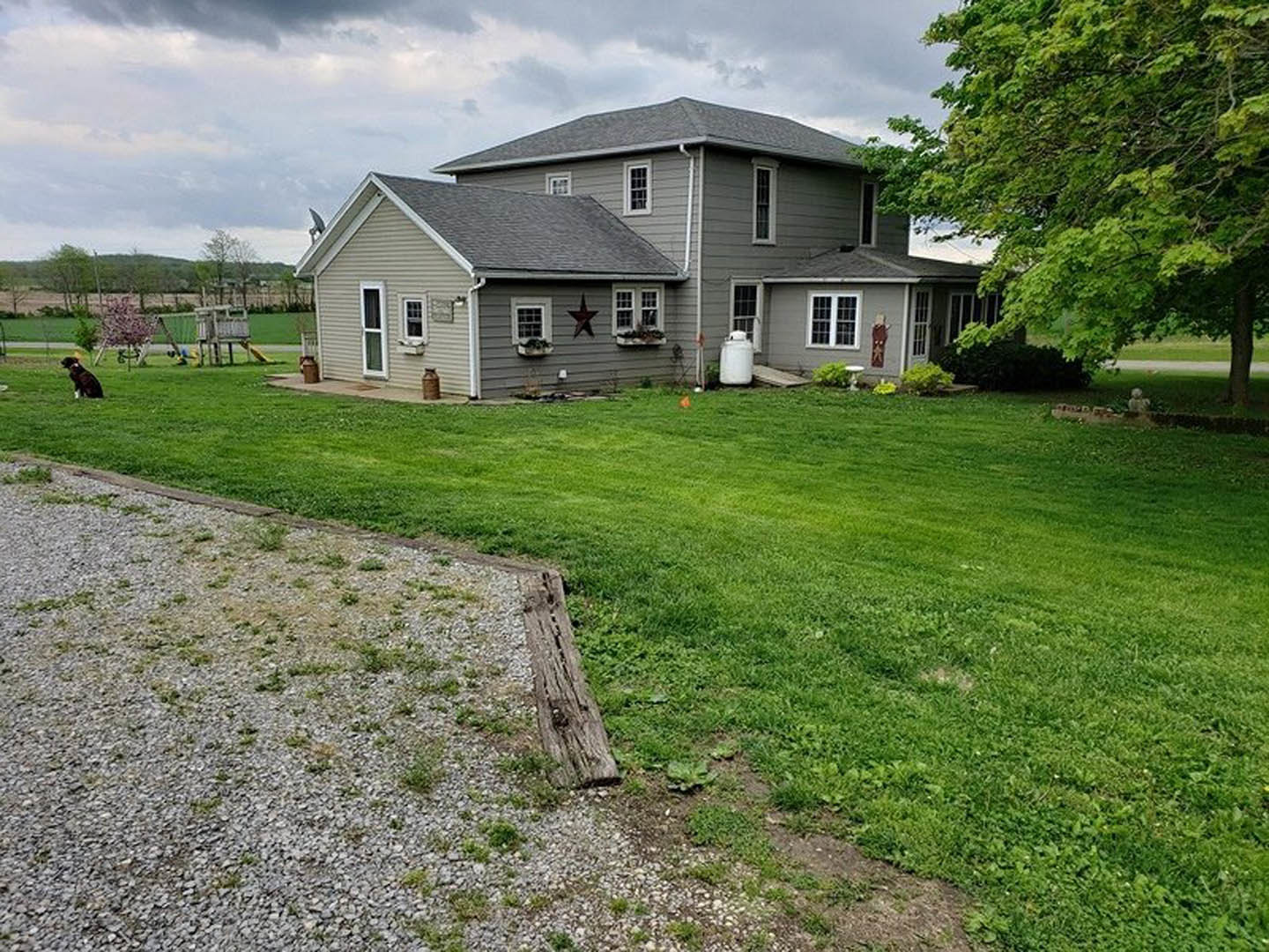 Grey farmhouse with star painted on siding, manicured lawn, tree in front yard, children’s playground set, cloudy sky overhead