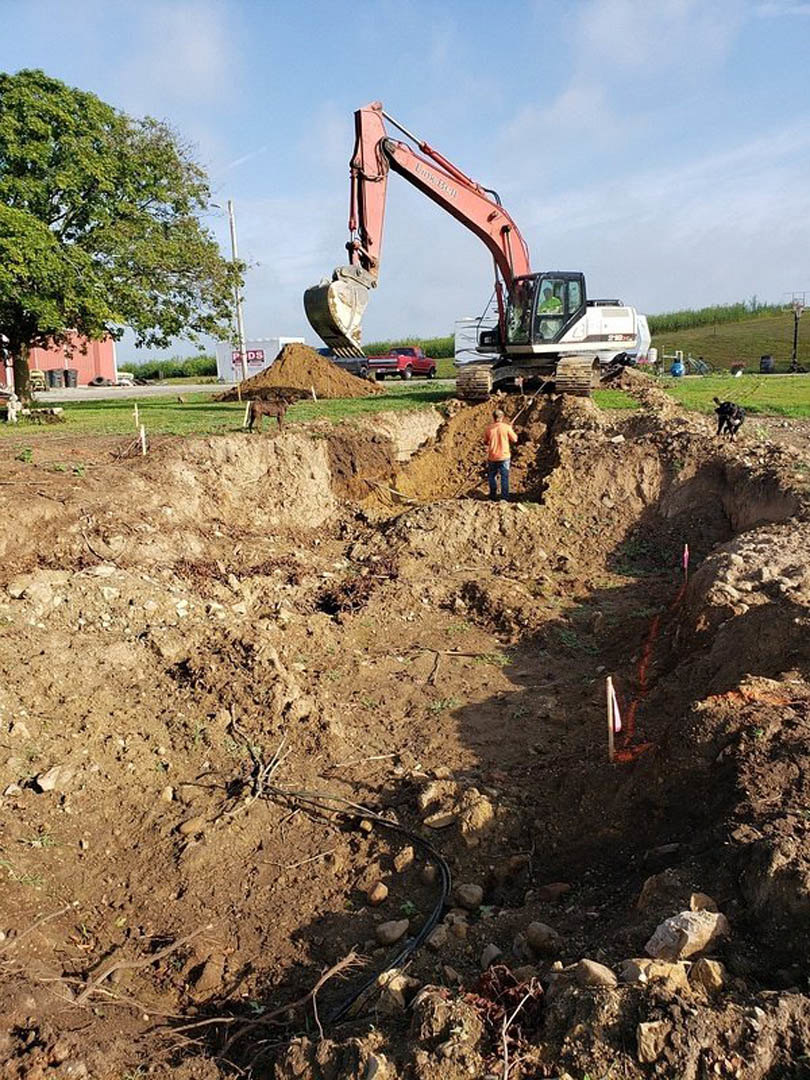 Excavator digging foundation pit on residential construction site with exposed soil, green-leaved tree in background, worker standing near machinery, scattered rocks and earth