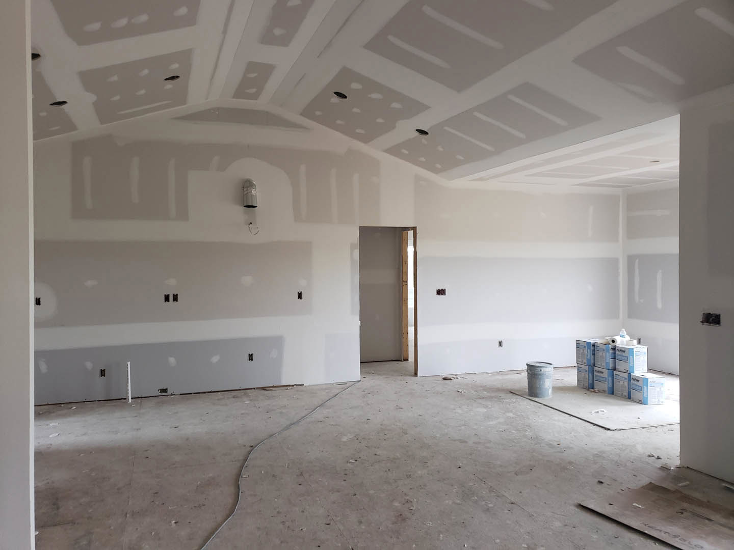 White and gray painted wall, exposed wooden framing, concrete floor with stacked boxes and a gray barrel, white door, ceiling with visible holes