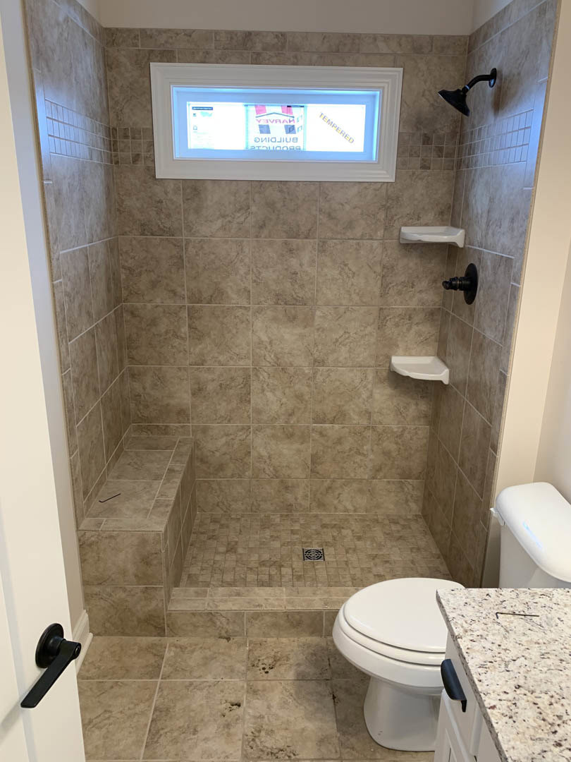 Modern bathroom featuring a glass-enclosed shower, large window with natural light, white tile walls, sleek countertop, black wall-mounted light fixture, chrome door handle, and