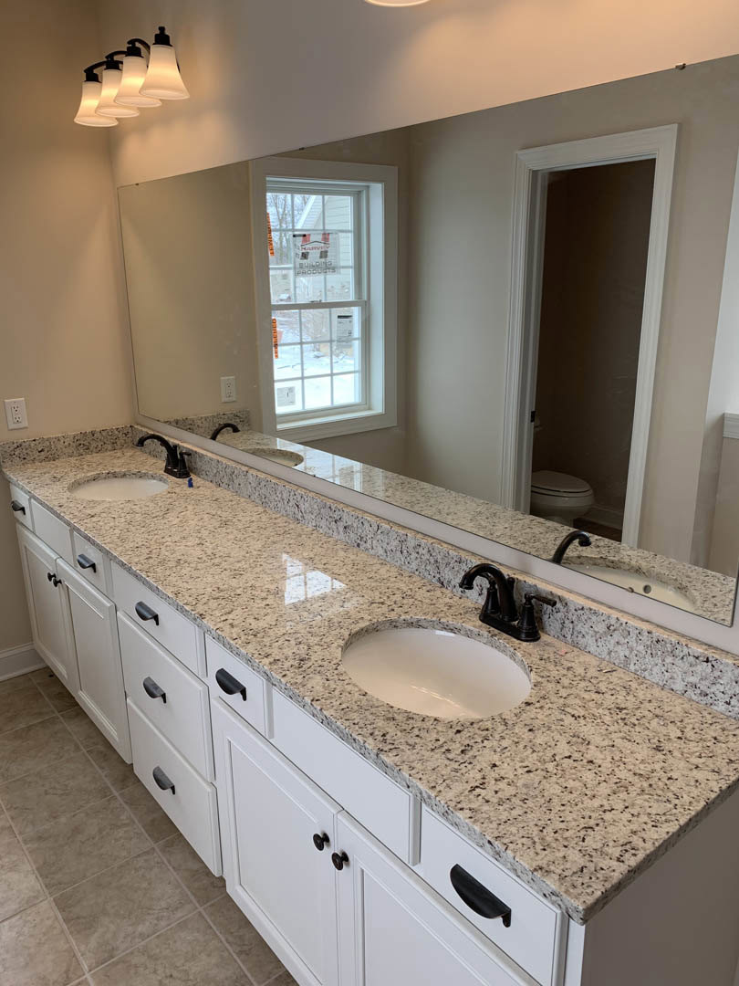 Bathroom with expansive mirror above double white sinks, black faucets, light fixture mounted overhead, open door frame, window with signage, white cabinetry and neutral
