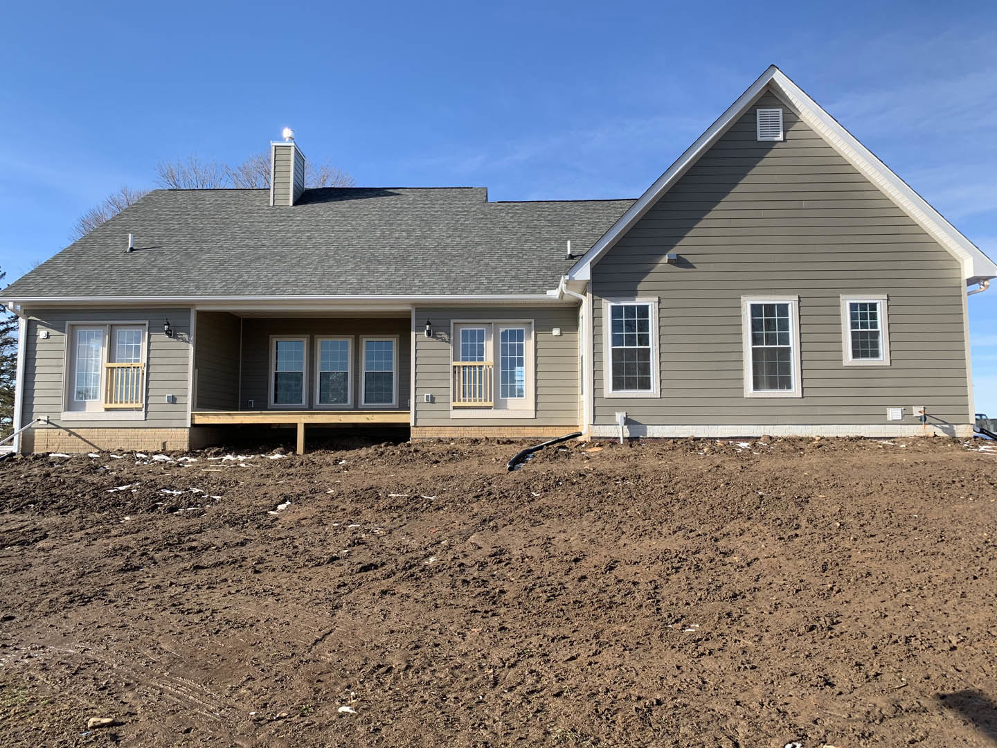 Two-story house with light siding, multi-pane windows, covered porch, and vent on exterior wall, set behind a dirt hill and field under a clear sky