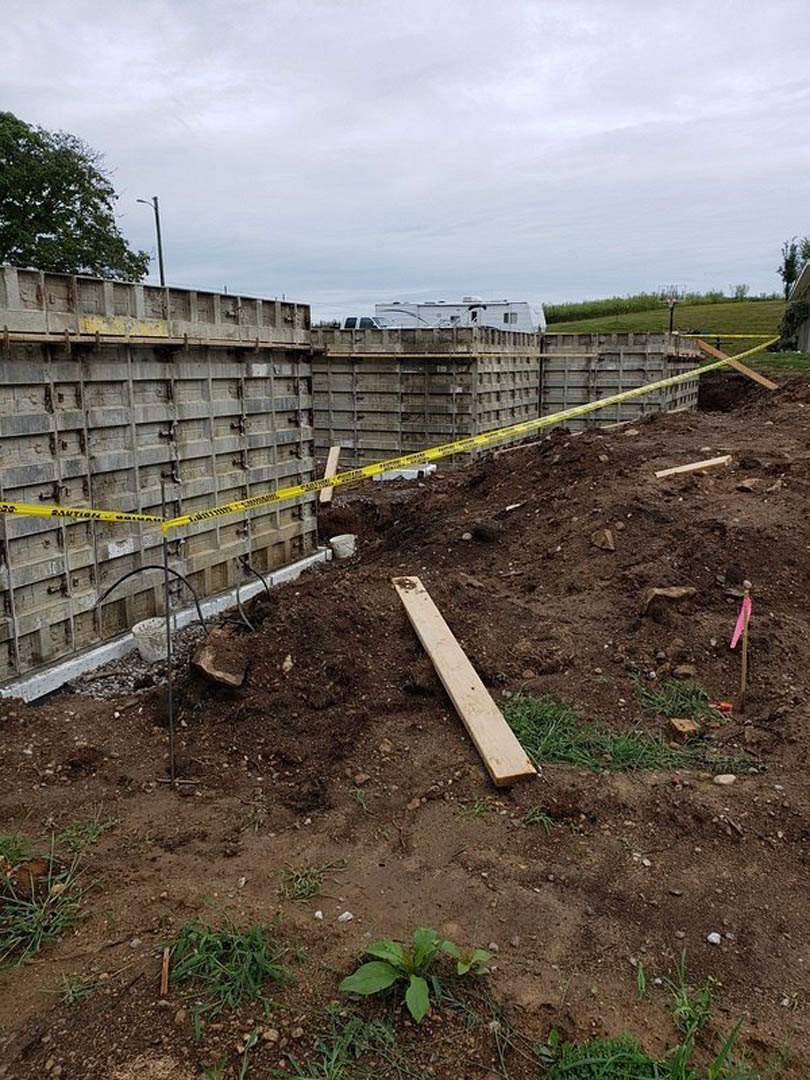 Wooden planks and yellow caution tape at a residential construction site, with a tree, green plants, and cloudy sky in the background.