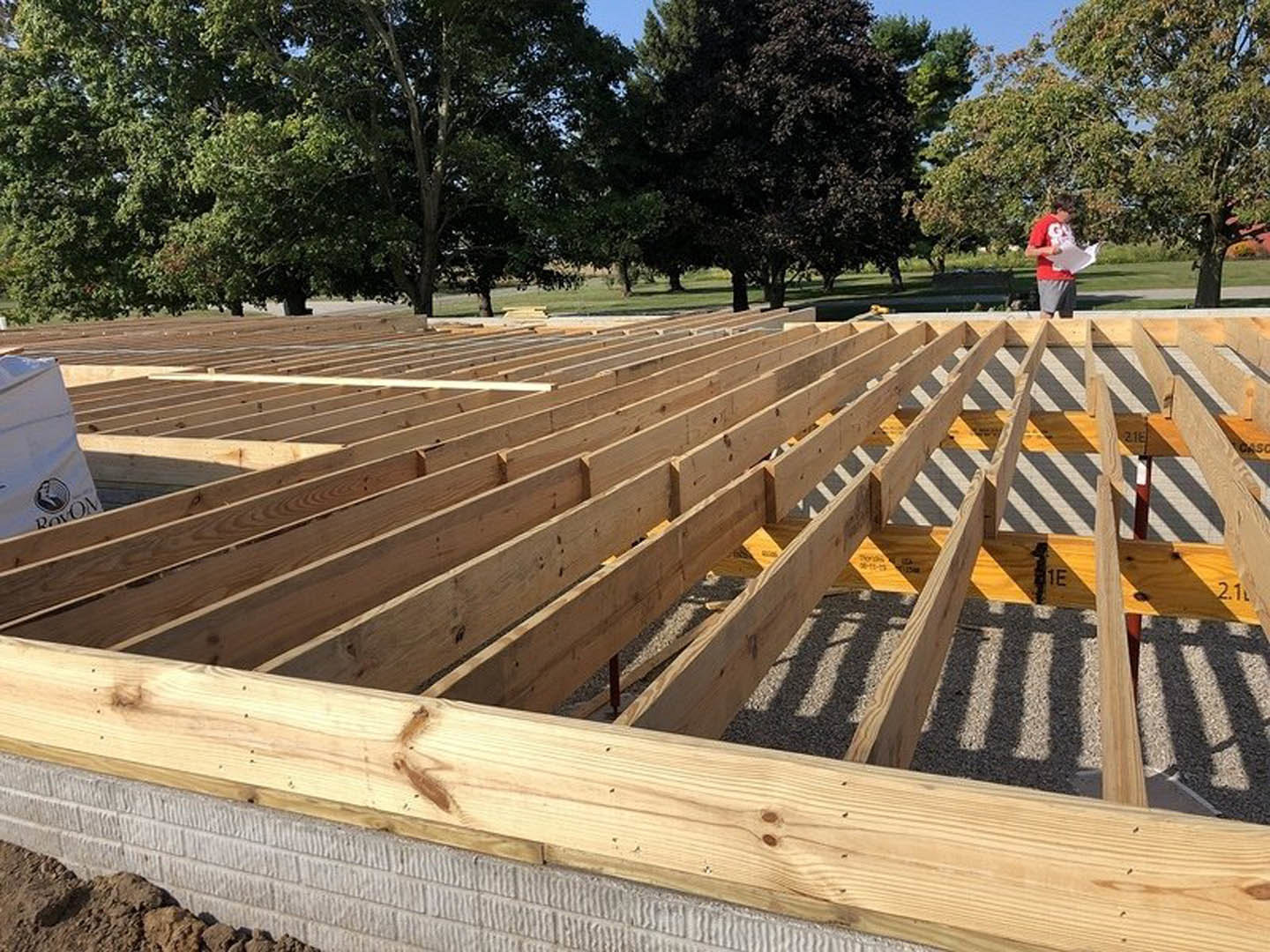 Wood-framed house under construction with exposed beams on concrete base, two people standing in front, surrounded by trees and outdoor materials