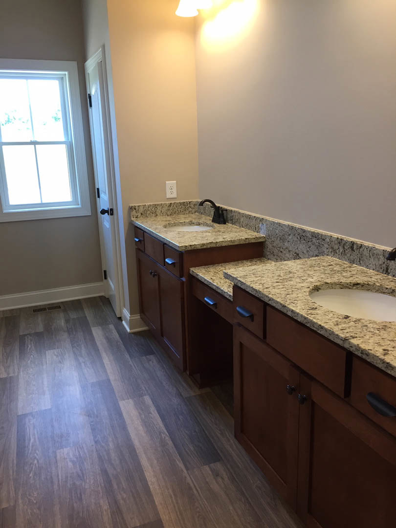Bathroom with dual sinks, marble countertop, wood flooring, white framed window, white cabinetry, and wall outlet visible.