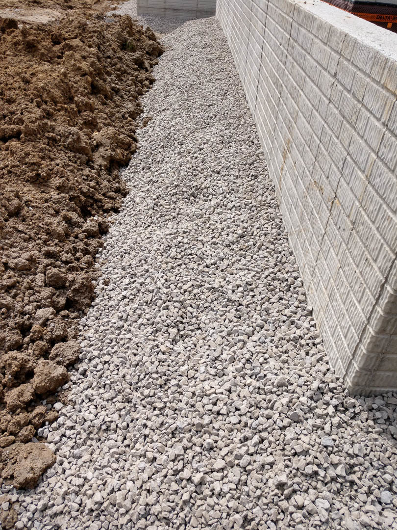 Stone exterior wall with gravel and dirt piled at the base, small rocks scattered on the ground