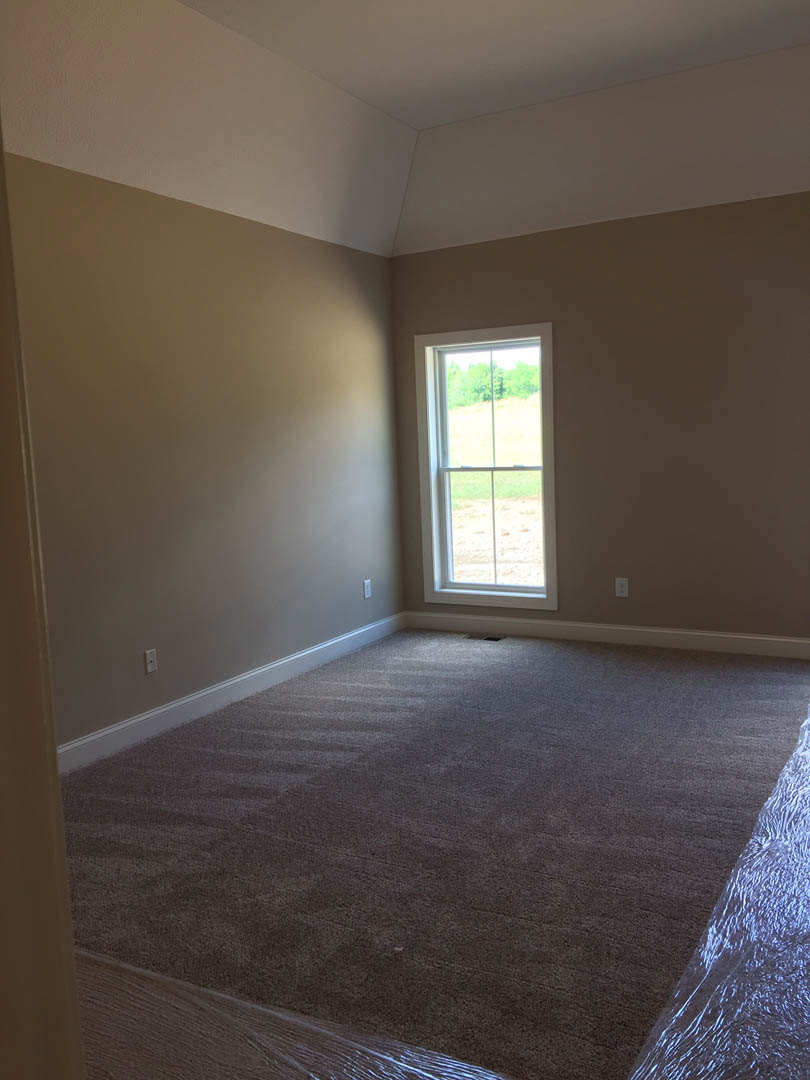 Carpeted bedroom with white walls, large window overlooking grassy field, baseboard trim, and natural light illuminating neutral finishes