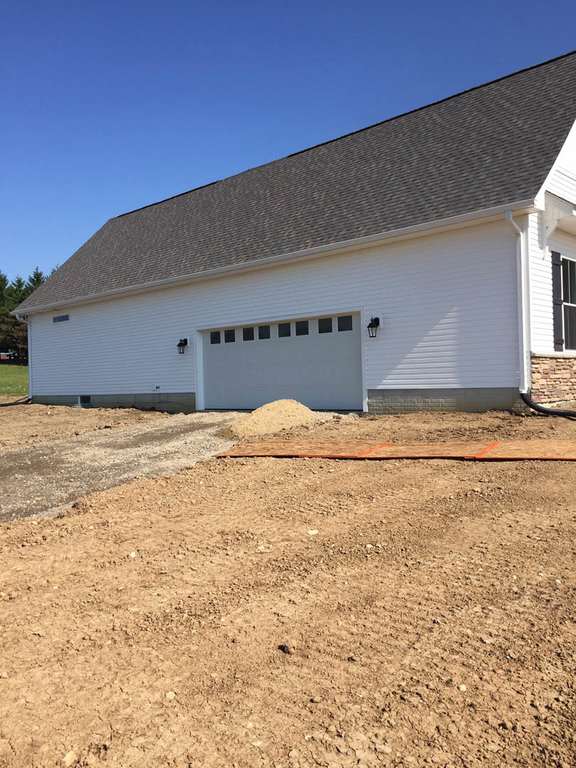 White house with horizontal siding, multiple windows, and a single garage door; dirt lot in foreground with scattered construction materials.