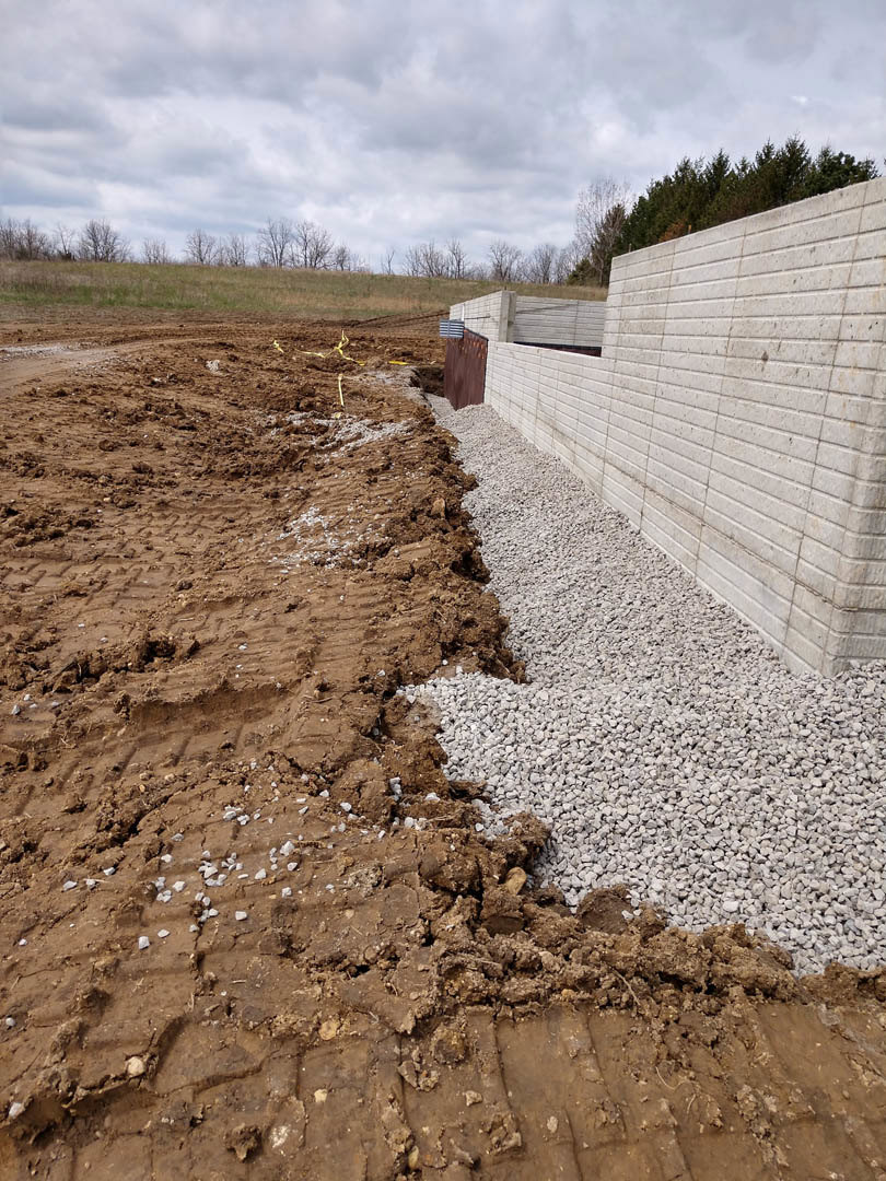 Gravel dirt road bordered by a brick wall, cloudy sky overhead, forested trees in background