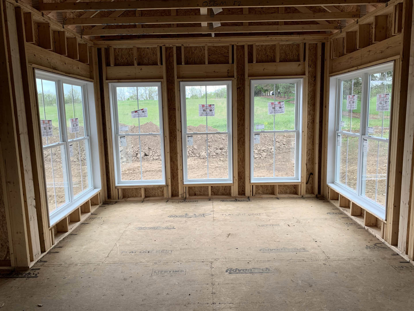 Spacious room featuring exposed wood ceiling beams, multiple large windows, light-colored walls, and a floor with black writing; green accent wall with a sign and a wooden shelf