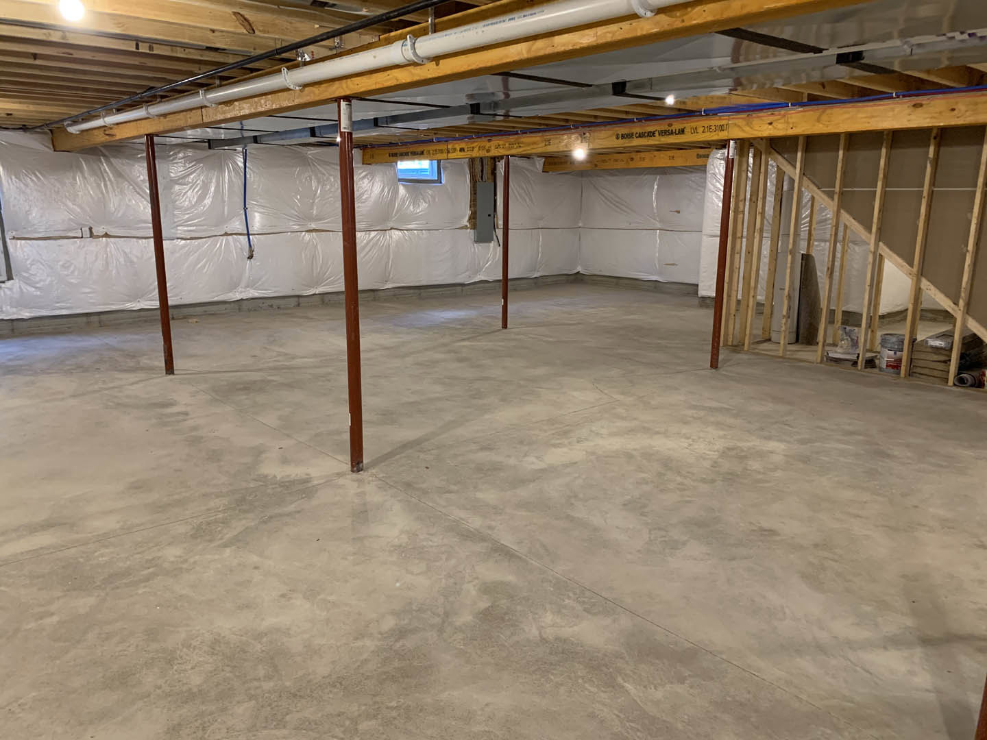 Concrete floor with exposed wooden post, white insulated wall, visible ceiling pipe and framing in unfinished basement room