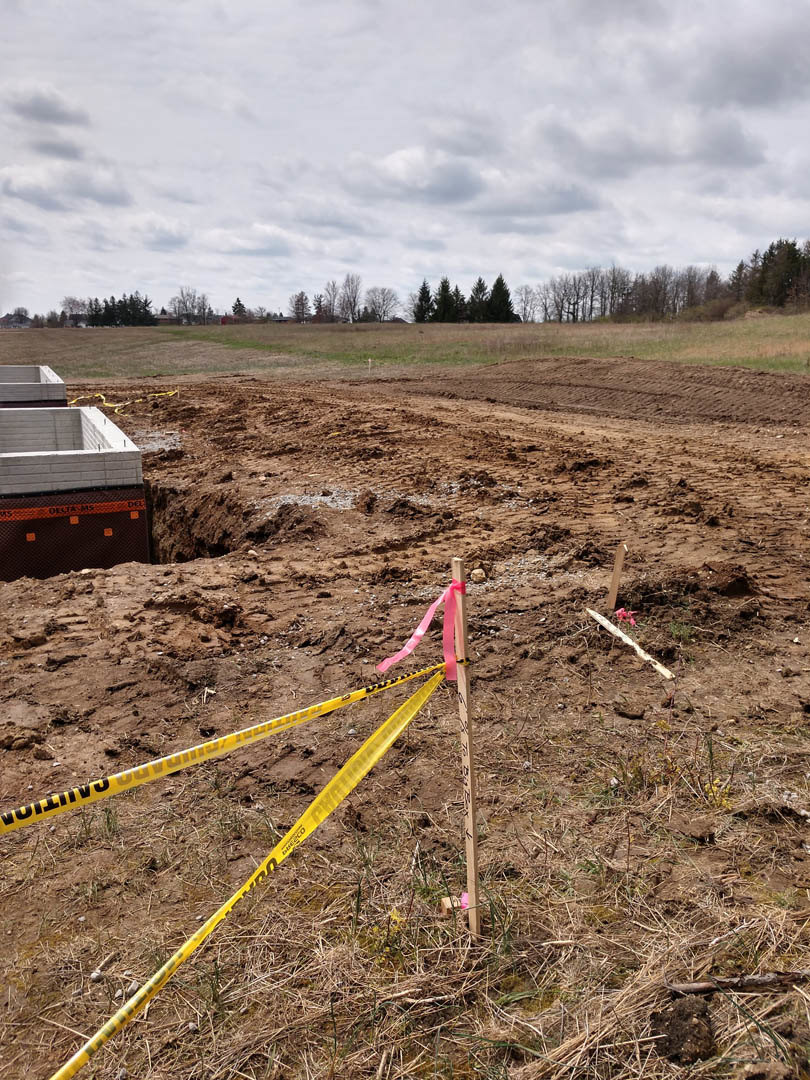 Foundation excavation surrounded by yellow caution tape, wooden posts, grassy field, cloudy sky, and distant trees