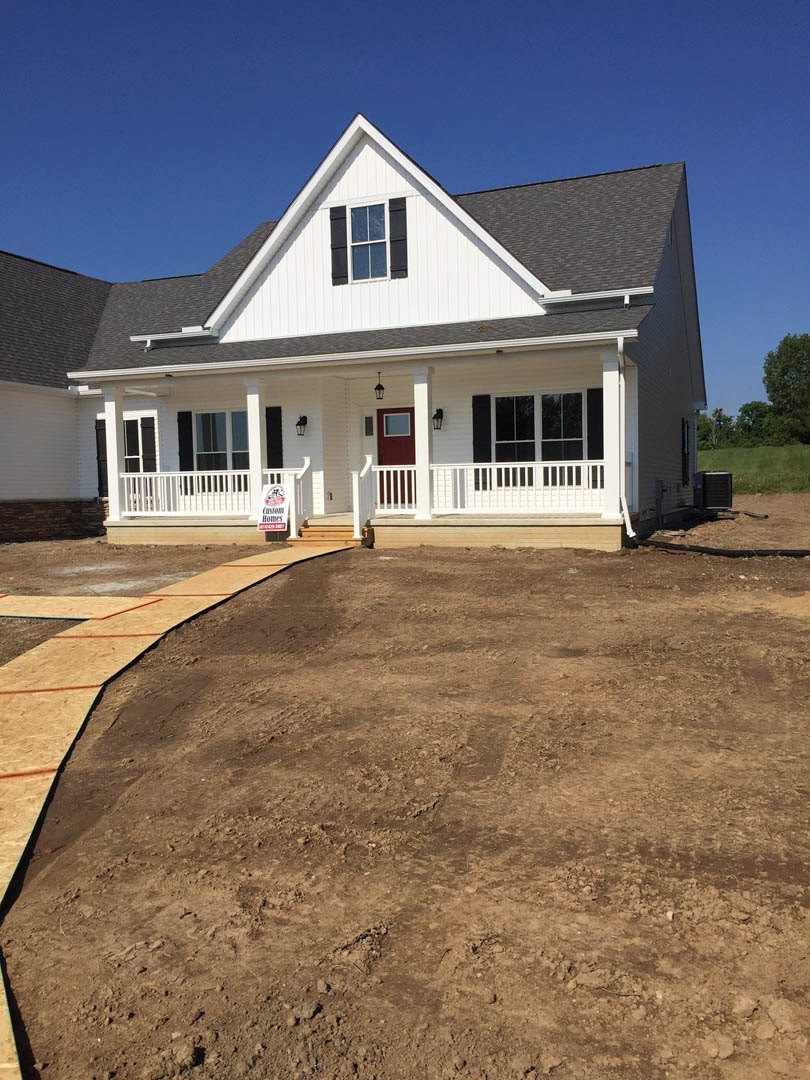 White house under construction with black shuttered windows, red front door, dirt path leading to porch, and trees in the background
