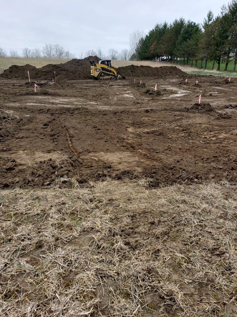 Yellow bulldozer with black roof parked on muddy dirt field, grassy ground and row of trees under blue sky in background
