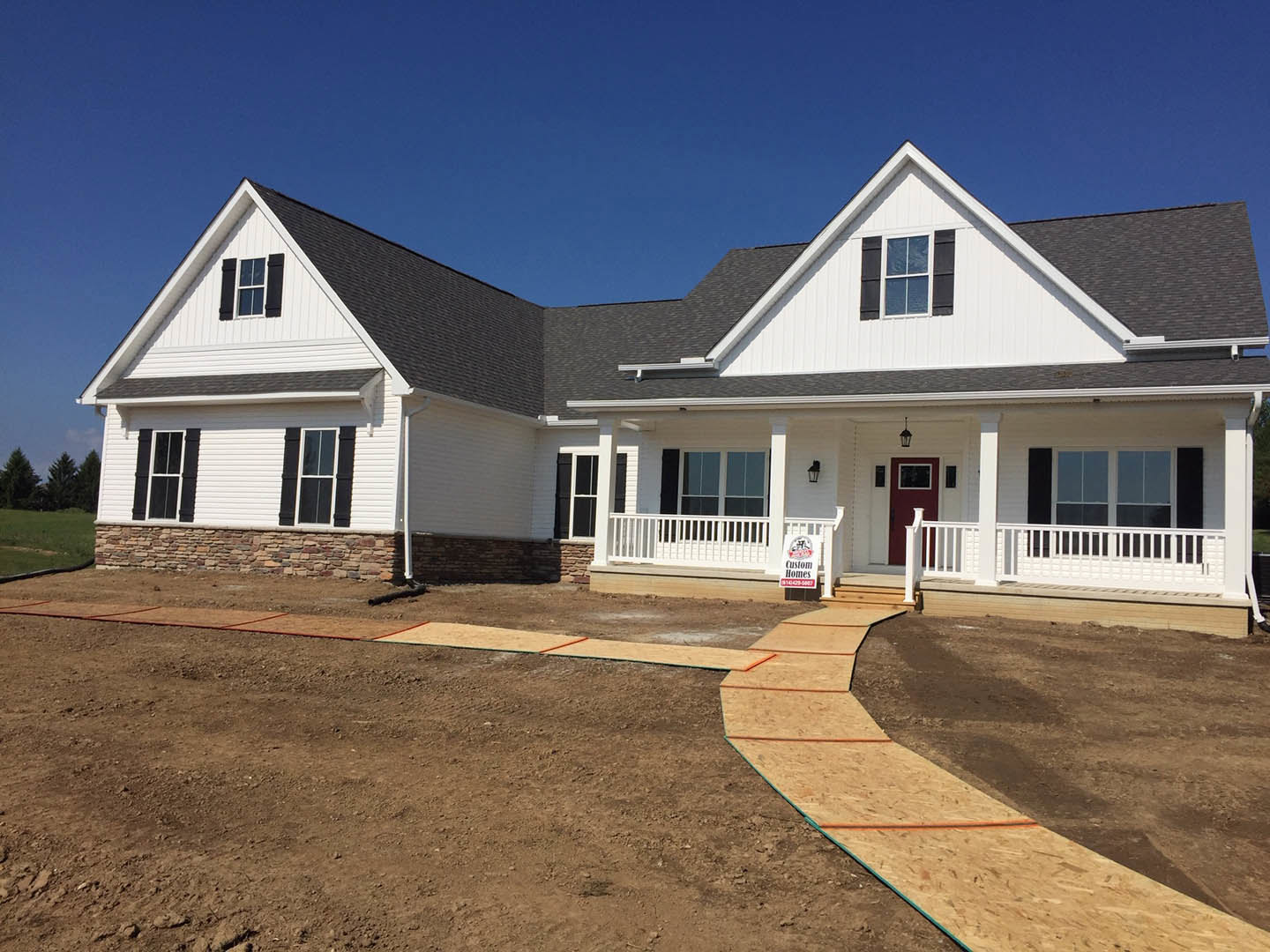 White siding house with black shuttered windows, red front door, white porch railing, paved driveway, and construction materials near brick wall.