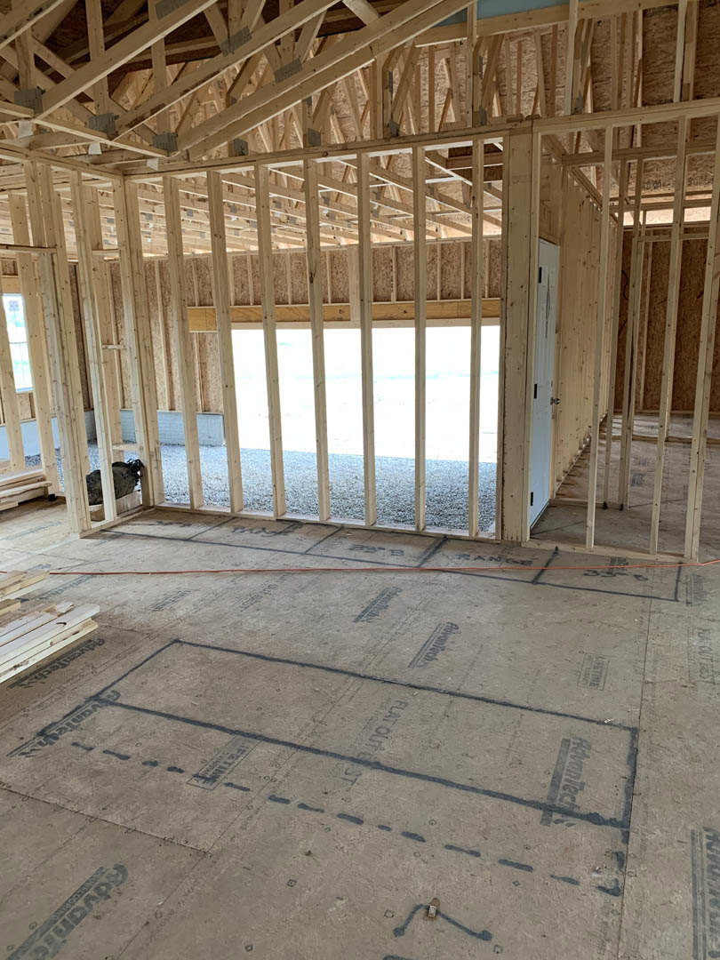 Unfinished room with exposed wood framing, subfloor marked with black lines, pile of wood planks, and a bird perched on a wooden ledge