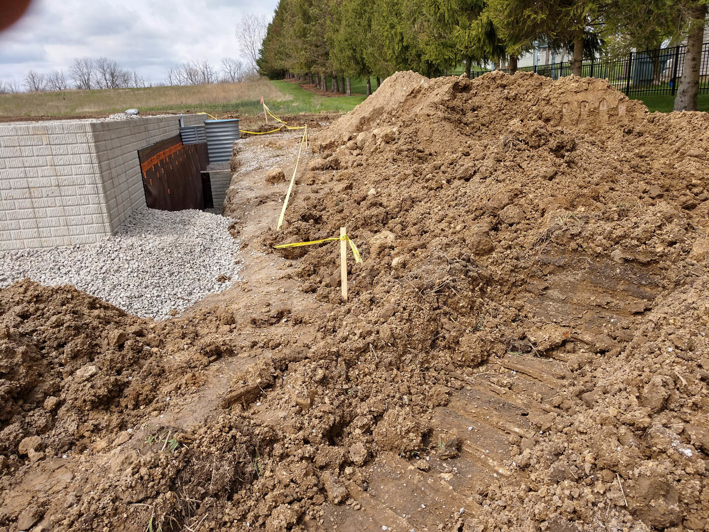 Mounded soil beside a red brick wall, with trees and plants in the background under an open sky