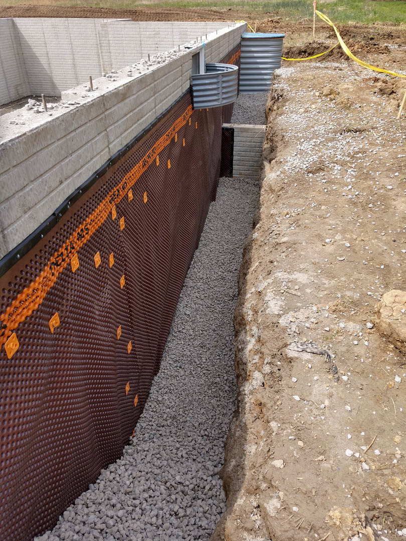 Concrete foundation slab with embedded drain pipe, yellow caution tape wrapped around metal pole, surrounding soil and patches of grass.